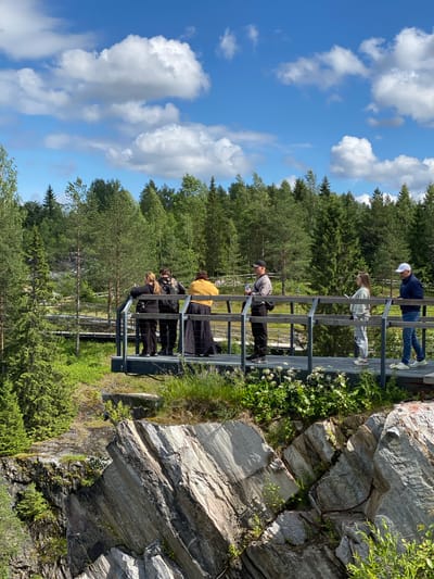 In the city of Ruskeala, Russia, a group of seven individuals stands on a metal viewing platform overlooking a rugged, rocky landscape and a dense forest under a partly cloudy sky. The platform, elevated above the terrain, features a metal railing.
The people are positioned at various points along the platform, engaged in observing the surroundings. From left to right, the first three individuals are close together, with two women and one person whose gender is not clearly discernible. One woman, wearing a black top and pants, is looking towards the cliff face. The person next to her, wearing a black jacket and pants, holds a bottle of water. The third person, a woman in a yellow cardigan and a long patterned skirt, is also looking out.
Further to the right, a man in a grey jacket and black cap holds a bottle of water. Next to him, a woman in a light-colored sweater and pants is looking at her phone. The last two individuals are a woman in a light-colored top and pants, and a man in a blue jacket and jeans, both looking in the same direction.
The environment consists of steep, layered rock formations in the foreground, with a dense evergreen forest covering the hills in the background. The platform is surrounded by green vegetation, including small white flowers along the edge. The sky is blue with scattered white clouds. The scene captures a moment of scenic appreciation in a natural, mountainous setting.
