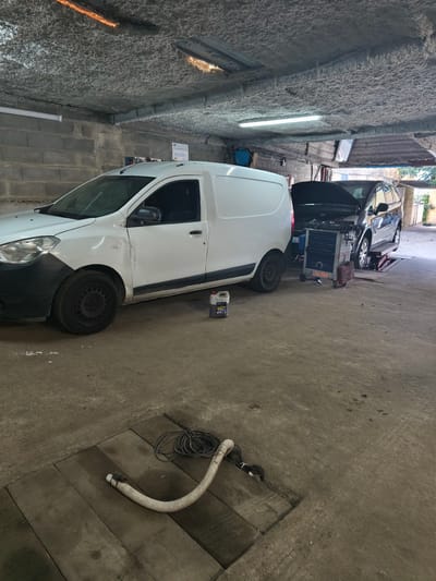 The image depicts the interior of an auto repair shop. A white cargo van is parked on the left. To the right of the van, a dark-colored car is on a lift, with its hood open. A mechanic's tool cart is positioned nearby. On the floor in the foreground, there are various cables and a corrugated hose. A bottle of oil is next to the van. The garage has cinderblock walls and a rough, textured ceiling. The location is Moaña, Spain.