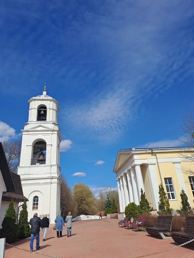 A low-angle shot, rotated counterclockwise, features two buildings under a blue sky with cirrus clouds. A white bell tower dominates the foreground, complete with visible bells and arched openings. Several individuals are present; some are walking near the tower, while others are seated on benches adjacent to a yellow building with white columns. The ground appears to be paved with red bricks, and landscaping includes trees and shrubs. A chain serves as a border. This is the Ivashevo, Russia.