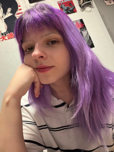 An indoor portrait photograph captures an individual with light skin, dark eyes, and long, straight bright purple hair with bangs, displaying darker roots at the scalp. The individual, seen from the chest up, wears a white polo shirt with horizontal black stripes. Their head is slightly tilted, and the right hand rests beneath the chin, supporting it. A subtle smile is present, and the gaze is directed towards the camera. The background is a light-colored wall featuring several posters: on the left, a black and white anime-style poster with Japanese text; above and to the right of this, a red poster with "VULGAR" text and a dark, contorted figure; further right, a dark vertical poster with white text, partially obscured. An unidentified dark, irregular object is visible in the top right corner. The specific geographical location in the city of Lubnya, Russia, cannot be determined from the visual information present in the image.