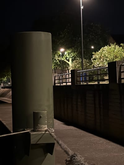 The image is a low-light, nighttime outdoor scene. In the foreground, a large, dark green, cylindrical metal object dominates the left side of the frame. Attached to the base of this cylinder is a smaller, upright cylindrical metal fixture with a dark circular indentation at the top, resembling an eye, and a rough, toothed band beneath it, possibly painted to look like teeth. A thick, gray rope is tied to this smaller fixture and extends to the right, trailing across a dark, textured surface which appears to be ground or a deck.

In the midground, a horizontal metal railing with vertical supports runs along the right side of the frame, separating an elevated walkway from the area in the foreground. Behind the railing, lush green foliage and trees are illuminated by artificial lights, creating bright patches. A dark, rectangular structure, possibly a building, is partially visible behind the foliage on the far right. A tall, dark lamppost with a light fixture at the top stands centrally in the background, casting illumination. The overall environment suggests an urban or semi-urban setting, possibly a waterfront or park area at night. There are no people visible in the image.

The image does not contain sufficient information to determine the specific location in the city of Charnay-lès-Mâcon, France.