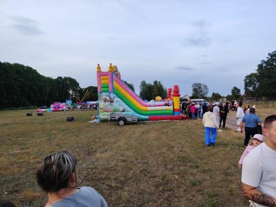 The image depicts an outdoor gathering in Олтушскі сельскі Савет, Belarus. In the center of the frame, a large, colorful inflatable slide with a rainbow design is set up. Several people are gathered around it, with some appearing to be waiting or observing. To the left, other inflatable attractions, including a pink and purple bouncy castle, are visible. A trailer is attached to the side of the large slide. The ground is covered in dry grass, suggesting a rural or park-like setting. Trees line the background. The sky is overcast with a pale gray hue. In the foreground, the backs of several people are visible, looking towards the event.