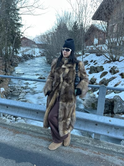 A single individual is observed standing on a road or bridge in Saanen, Switzerland, during a winter season. The person, appearing female, is positioned centrally, facing slightly right and looking away from the camera. She wears a black knit beanie, white-framed wraparound sunglasses, and a long, multi-toned brown faux fur coat. Underneath, she has dark brown wide-leg pants and tan UGG-style boots. Black gloves are worn on both hands. A small black shoulder bag hangs over her left shoulder, and a light green smartphone is held in her gloved left hand. She is leaning against a grey metal guardrail that runs horizontally across the lower third of the frame.
To the left and right of the subject, the ground is covered in snow. A river or stream flows behind the guardrail, featuring moving water and visible ice formations, particularly around rocks and edges. Bare deciduous trees and some evergreen conifers line the riverbanks. In the background, traditional wooden chalets with sloped roofs are visible through the trees on both sides of the river, nestled into the snowy landscape. The sky is overcast and light-colored. The immediate foreground shows a dark grey paved surface.