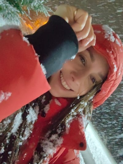 The image depicts a female individual outdoors in Dolmatovo, Russia, under snowy, low-light conditions. She is positioned at an angle, facing the viewer, and smiling with visible teeth. Her dark, medium-length hair and the hood of her red puffer jacket are dusted with snow. A black long-sleeved garment is visible at her right wrist. Her right hand, adorned with a gold-colored ring on the ring finger, is raised with fingers curled, resting near her forehead. In the blurred background to the upper left, snow-covered green coniferous tree branches are visible, behind which an orange light source glows. The background on the right consists of dark, indistinct areas and a snow-covered surface at the bottom right.