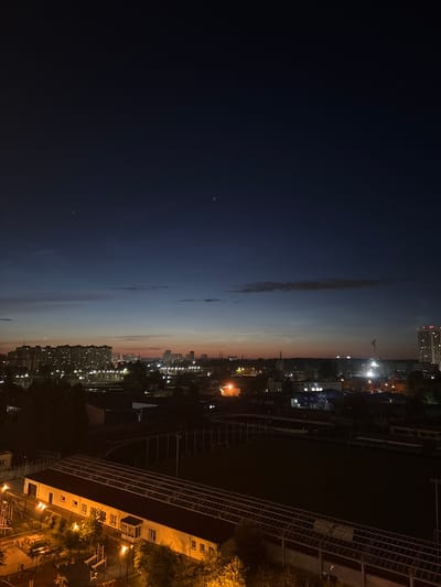 This is an aerial view of a city at dusk or night. The left side of the image is dominated by a deep blue, starry sky, transitioning to lighter shades of blue and then a warm, orange-pink hue as it meets the horizon. A long, thin cloud stretches across the sky in the middle. The right side of the image shows a cityscape with illuminated buildings and streetlights. A large, dark building with a grid-like structure, possibly a stadium or industrial facility, occupies the foreground on the right. Trees and smaller buildings are visible behind it. The overall impression is one of urban life transitioning into night, with a hint of a soft sunset. No people are clearly visible. The time of day is indicated by the twilight sky and the artificial lights of the city. The location is identified as Lesnye Polyany, Russia. There is no visible text in the image.