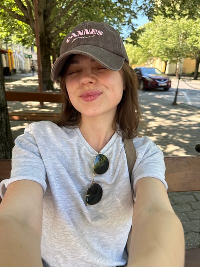 A selfie of a light-skinned young woman with brown hair sitting outdoors. She is wearing a grey baseball cap with "Cannes" in pink letters, a grey long-sleeved shirt, and sunglasses are hanging from her shirt. The woman is sitting on a bench, possibly near a street in Siófok, Hungary. The background includes trees, shadows, buildings, a car, and pedestrian traffic control barriers. The photograph is taken at an angle with a slight downward tilt.