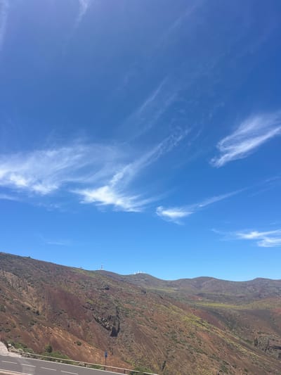 The image depicts a natural landscape under a bright blue sky with wispy white clouds. The foreground shows a paved road with white lines, bordered by a wooden fence and some low-lying vegetation. Beyond the road, a steep, reddish-brown hillside rises, characterized by varying shades of brown, green, and patches of sparse vegetation. The terrain is rugged and uneven, with visible erosion patterns and rocky outcrops. In the distance, several white structures, possibly scientific or communication towers, are visible atop the hills. The sky takes up a significant portion of the image and is clear except for the scattered clouds. The scene appears to be a mountainous or hilly area, possibly a volcanic region given the reddish soil. The image was taken in La Orotava, Spain.