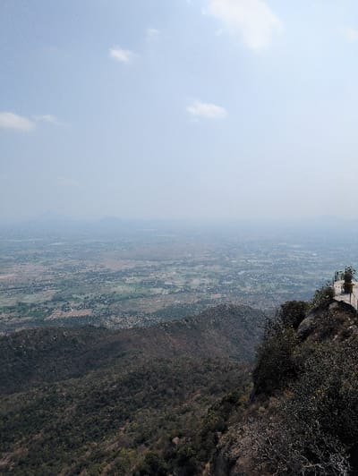 A panoramic view from a high, rocky, vegetated elevation in the city of Kadaladi, India, extends across a vast, hazy landscape under a pale blue sky with scattered white cumulus clouds. In the immediate foreground on the right, a portion of a man-made structure is visible, featuring a light-colored railing consistent with a viewing platform, surrounded by dense, dark green to brown scrub vegetation and exposed rock. The steep hillside below is heavily covered with similar dark foliage. Beyond this, a wide valley or plain stretches into the far distance, presenting a mosaic of green agricultural fields, lighter brown patches of land, and numerous small clusters of buildings indicative of human settlements. Distant mountain ranges are faintly discernible on the horizon, partially obscured by atmospheric haze. No human figures, objects, or specific actions are observed within the visible scene.