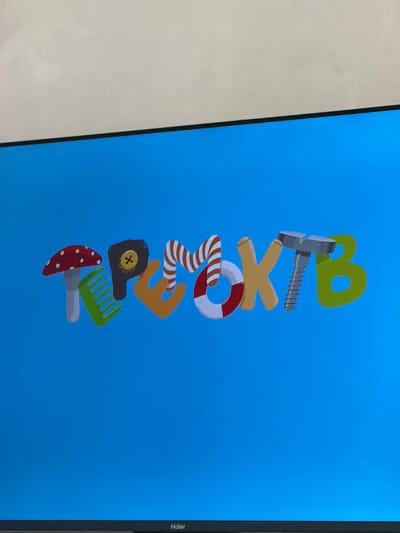 The image shows a television screen displaying the word "ТРЕОЖТВ" in stylized, colorful letters. The letters are designed to look like various objects: 'Т' resembles a mushroom, 'Р' is a green comb, 'Е' is a fuzzy brown object with a yellow button, 'О' is a life preserver, 'Ж' is a light brown letter with stitching, 'Т' is a silver screw, and 'В' is a green letter. The background of the screen is a solid blue color. The bottom of the television screen has a black bezel with the word "Haier" printed on it. The context of Krasnyi Krym, Russia is not visually represented in the image.