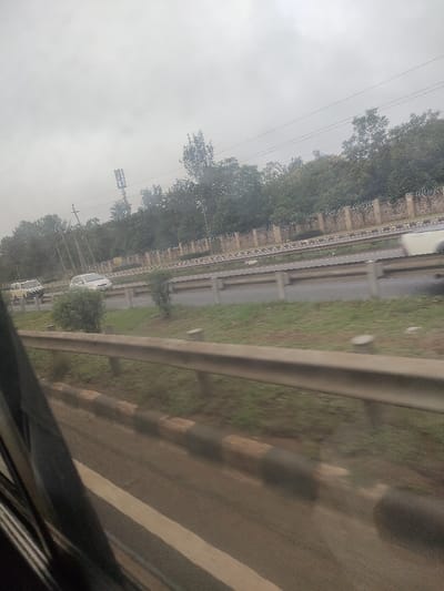The image captures a view from a moving vehicle, looking towards a multi-lane highway on an overcast day. In the immediate foreground, a dark, blurry section of a vehicle's interior is visible on the left, with a horizontal silver guardrail spanning the lower mid-section. Beneath the guardrail is a dirt shoulder adjacent to a curb painted with alternating black and white stripes.
The midground features a paved highway with two visible lanes of traffic. A grassy median strip with small bushes separates the lanes. In the leftmost lane, a white sedan is in motion, followed by a yellow-and-white striped vehicle, possibly a commercial van or matatu. A blurry white vehicle, consistent with a pickup truck or SUV, is partially visible and in rapid motion in the rightmost lane.
In the background, a decorative stone or brick wall, possibly topped with barbed wire or security fencing, runs parallel to the highway. Behind this wall, a dense line of green trees forms a natural barrier. Above the treeline on the left, a multi-mast cellular tower stands prominently, accompanied by utility poles and overhead power lines. The sky is uniformly grey, indicating overcast conditions.
No specific geographical markers are present within the image to definitively identify the location as Gatongora ward, Kenya. No people are explicitly visible or identifiable in the scene.