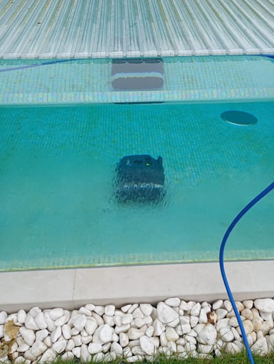 The image depicts a swimming pool with a robotic pool cleaner operating underwater. The pool is filled with clear, turquoise water and has a tiled bottom. A blue hose is visible, presumably connected to the pool cleaner. The edge of the pool is lined with white stones and green grass. In the background, a translucent pool cover is partially visible. The scene is clean and well-maintained, suggesting a residential or commercial setting. The location is identified as Nowa Wieś Szlachecka, Poland.