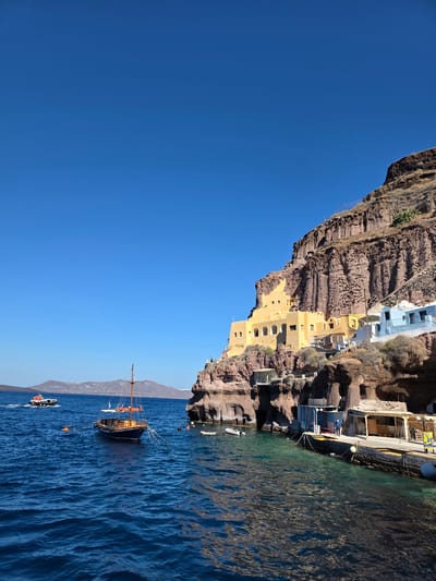 An eye-level, wide shot captures a bright, sunny day at the port of Ammoudi Bay, located below Oia in the Thira Municipal Unit, Greece. The deep blue sea fills the lower half of the frame, with gentle waves reflecting the clear sky. In the middle ground, a traditional wooden sailboat with an orange sail is moored. To its left, a white and red boat, possibly a water taxi, cruises away from the shore. The steep, rocky cliffs of the caldera rise dramatically on the right, dotted with traditional Cycladic architecture. A cluster of yellow buildings, featuring arched windows and balconies, cling to the cliff face. Below these, a light blue building with white trim is visible. At the base of the cliffs, a small pier area features a few structures, including a covered seating area and a person standing near a railing. The scene is tranquil, with no other people visibly interacting. The overall impression is one of picturesque coastal beauty.