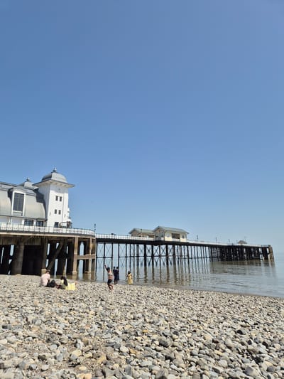 The image shows Penarth Pier in Penarth, United Kingdom, taken on a bright, sunny day. The pier extends from the lower left diagonally to the upper right of the image. The pier structure is visible, including support beams and building segments. The pier is constructed with dark metal supports. The photograph is from a low angle, depicting a pebble beach in the foreground, and the pier extending over a calm sea. Several people are present on the beach and under the pier. Some are seen playing or relaxing near the water's edge. The environment is characterized by clear blue skies. The photograph is taken in a vertical orientation.