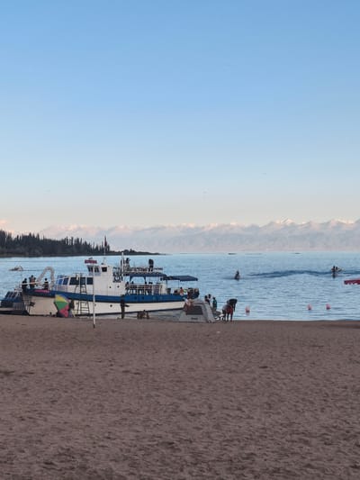 This image depicts a serene beach scene on the shores of Issyk-Kul Lake, near the city of Bayet, Kyrgyzstan. In the foreground, a sandy beach stretches out, with a few scattered objects and people. A large white boat with blue trim is moored near the shore, with several individuals visible on its deck and the surrounding area. One person stands on the beach next to the boat. Colorful beach umbrellas are visible, one of which is set up on the sand.
The middle ground features the calm, blue waters of Issyk-Kul Lake. Several people are enjoying water activities; some are on jet skis, creating wakes in the water, while others appear to be swimming or wading. Buoys are scattered in the lake, marking a designated swimming area.
In the background, a dense line of dark green coniferous trees borders the lake, backed by a majestic range of snow-capped mountains that stretch across the horizon. The sky is a clear, light blue, indicative of fair weather. The overall atmosphere is one of leisure and natural beauty.