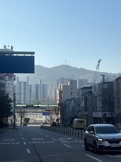 An urban landscape features buildings of varying heights and architectural styles along a road. A distant mountain is partly obscured by haze. A construction crane rises above the structures. Vehicles, including cars and a yellow truck, are visible on the right side of the street. A large blue billboard stands to the left. Overhead power lines run along the skyline.