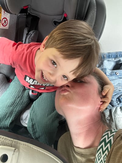 A photograph taken from a high angle shows a child and an adult, likely in a vehicle. The child is in a car seat, wearing a red t-shirt with white and black lettering. The child is looking up and smiling at the camera. The adult is leaning in, with their face close to the child's face, as if giving a kiss. The adult is wearing a green top with a white geometric pattern. The seat in the background is gray, and there's a blue denim jacket or shirt. The car's interior is visible. It is not possible to ascertain the location as Meljine, Montenegro, from this image.