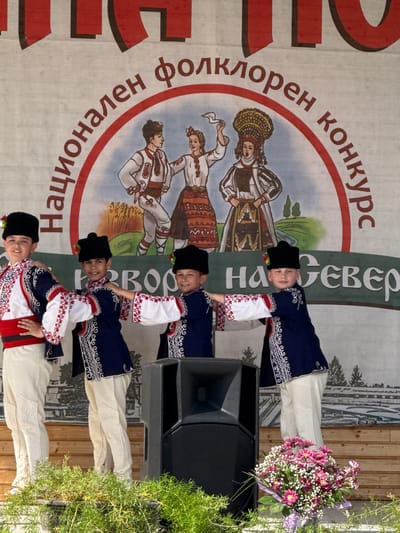The image depicts a group of four young boys in traditional Bulgarian folk costumes performing on an outdoor stage. They are positioned in a line, facing forward, with their arms outstretched and hands placed on the shoulders of the person in front of them. All four boys are wearing dark, wide-brimmed hats adorned with small red and yellow flowers, and embroidered dark blue vests over white shirts with intricate red and white embroidery on the sleeves. They also wear cream-colored trousers with decorative stitching.
Behind the performers, a large banner is visible, featuring a circular illustration of people in traditional attire and text in Cyrillic script. The text, partly obscured by the performers and stage equipment, appears to be "Национален фолклорен конкурс" (National Folklore Competition). The banner also has a dark green horizontal band across the bottom with more Cyrillic text, likely indicating the location or name of the event.
In the foreground, a large black speaker is placed on the stage to the right of the performers. To the left of the speaker and in front of the stage, a vibrant bouquet of pink and white flowers is arranged. The stage itself is made of wooden planks, and the background suggests an outdoor setting with greenery and possibly trees. The overall atmosphere is festive and suggests a cultural performance.
This event is likely the National Folklore Competition held in Pordim, Bulgaria, as indicated by the text on the banner.