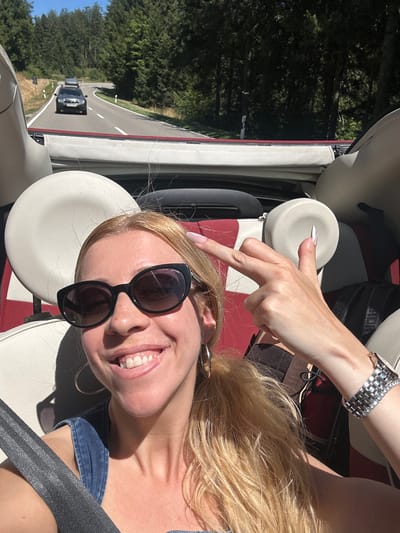 A woman with blonde hair and sunglasses is in the front passenger seat of a convertible, smiling and giving the peace sign. She is wearing a denim outfit and a watch. The car is on a road in a forested area, with other cars visible in the distance. The scene appears to be in or around Neukirch, Germany, indicated by the road signage.