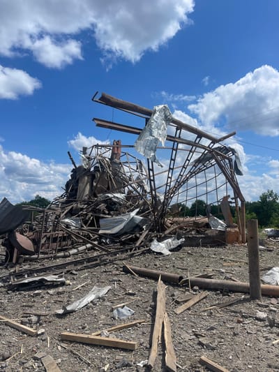 The image shows the aftermath of a destruction in Terny, Ukraine. The scene is characterized by the remnants of a metal structure, likely a building, destroyed and scattered across the ground. The structure, originally composed of metal beams, now lies in a jumbled heap of debris. Sheet metal fragments, twisted and torn, are strewn among the wreckage. Some wooden beams are visible, contributing to the overall sense of disarray. The ground is covered in debris, a mixture of metal, wood, and dust, indicating the severity of the destruction. The sky is blue, partially obscured by white, fluffy clouds, providing a backdrop for the somber scene. No people are present in the image.