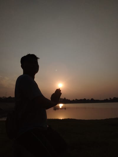 A silhouette of a person is seen from the side, facing a body of water at dusk. The person is kneeling with their hands pressed together in front of their chest, in a prayer or meditation gesture. The setting sun casts a warm glow on the scene, reflecting on the water. The background shows a distant treeline and a few structures, suggesting a suburban or rural area. The location is identified as Blimbing, Indonesia. The sky is a gradient of pale orange and gray, indicating the end of the day. The overall mood is peaceful and serene.