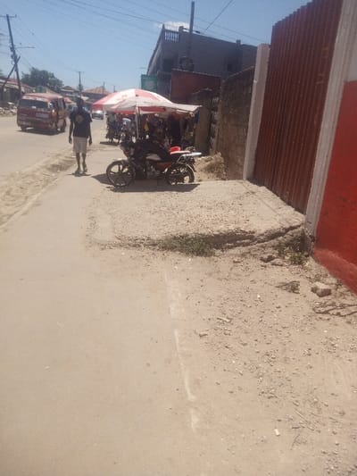 This image depicts a daytime street scene in Mtopanga ward, Kenya. The foreground shows a light-colored, possibly asphalt or compacted dirt road surface extending from the bottom left, with a faint white line marking its right edge. To the right of the road is an unpaved, uneven dirt and gravel shoulder, interspersed with sparse green vegetation and concrete sections.
On the paved road in the mid-ground, a male individual, wearing a dark t-shirt and light shorts, walks away from the viewer. Further down the road, a red van or public transport vehicle is partially visible, also moving away.
To the right of the walking individual, on the dirt shoulder, a black and red motorcycle is parked facing left. Immediately behind the motorcycle, a red and white striped market umbrella provides shade over an area with several indistinct figures and what appears to be merchandise, suggesting a street vendor setup.
Behind the market stall, a multi-story building with a dark facade and a rooftop structure is visible. Overhead power lines span the clear blue sky.
The far right of the image features a tall, vertical structure, possibly a gate or fence, constructed from red corrugated metal panels with white trim. Adjacent to this, a rough-textured, possibly stone or concrete wall extends towards the mid-ground, creating a boundary between the dirt shoulder and what appears to be another property.