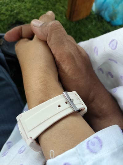 The image shows a close-up of two people holding hands. One person's hand is darker skinned and appears to be larger, while the other person's hand is lighter skinned and has a pale pink watch on their wrist. The lighter-skinned person's arm is resting on a white blanket with purple circular patterns. The background is blurred, but appears to be outdoors with green grass and possibly furniture. The scene suggests a moment of connection and support between two individuals. The lighting indicates it is daytime. The location context of Keudekruenggeukueh, Indonesia, is provided but not visually evident in the image itself. No visible text is present in the image.