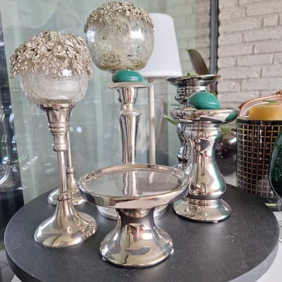 This media file features a close-up, horizontal shot of three ornate silver-plated candlesticks with teal accents, placed on a reflective surface, likely a table. The candlesticks are positioned from front to back, with the closest one being the most detailed. It has a round, textured silver base adorned with intricate floral and leafy patterns encrusted with small, clear crystals. Above this decorative base is a ribbed silver stem, followed by a teal gemstone, and then a fluted silver cup designed to hold a candle.
The second candlestick, slightly behind and to the left, shares a similar design with a teal gemstone and a ribbed silver stem, but its base is less ornate, appearing smoother. The third candlestick, mostly obscured by the others, shows a glimpse of its silver stem and a teal accent.
Behind the candlesticks, a white planter with a green leaf plant is visible. Further in the background, a wall with a textured, light-colored brick pattern and a decorative dark green and gold object, possibly a vase or lantern, are present. The overall setting appears to be indoors, possibly a home or a shop display, with soft lighting. There is no discernible text or information about the time of day, weather, or specific emotions conveyed. The focus is solely on the decorative objects.
