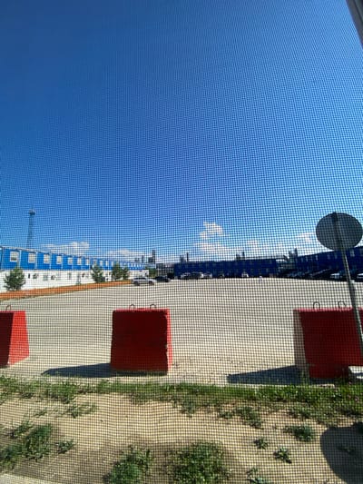 This image shows a view through a screen of a blue and white industrial building under a clear blue sky with sparse clouds. In the foreground, there are three large red concrete blocks, two of which are fully visible and one partially visible on the far left. A paved area is visible behind the concrete blocks, and a parking lot with several cars is situated in front of the building. Trees are planted along the perimeter of the building. A circular traffic sign is visible on the right side of the image. The scene appears to be an industrial area in Yukhta, Russia, on a sunny day.