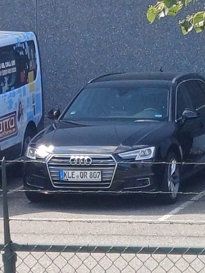 A black Audi A4 Avant wagon is parked on asphalt. The vehicle's front license plate displays "KLE QR 807" and is blue and white, indicating it is from Germany. The car is parked next to a white and blue van. The van has writing on its side, including "OTTO" and what appears to be a web address. The scene is outdoors, with a grey, textured wall in the background and a chain-link fence in the foreground. Green leaves from a tree are visible at the top right of the image. The image appears to have been taken in Venlo, Netherlands, based on the license plate format and surrounding context.