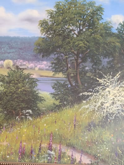 The image is a painting of a pastoral landscape. In the foreground, a path winds through a field of wildflowers. The wildflowers are depicted in various shades of purple, yellow, and white, with tall green grasses interspersed among them. To the right of the path, a large bush or tree is covered in white blossoms. In the mid-ground, a river flows gently, reflecting the sky. On the far bank of the river, there are rolling hills covered in trees. In the distance, a hazy sky with scattered clouds completes the scene. The painting evokes a sense of peace and tranquility. There are no people or animals depicted in the painting. The location in the city of Lyepyel, Belarus is not identifiable from the image.