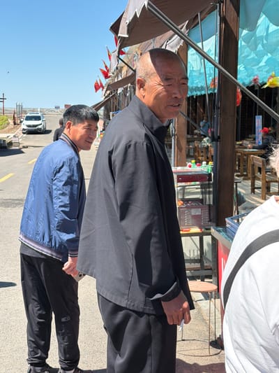 This outdoor photograph depicts a bustling market street in Jiejinkou, China, under a bright, clear blue sky. In the foreground, an older man with a shaved head and a pleasant expression looks upwards and to his right. He is wearing a dark, long-sleeved jacket or shirt and holds a lit cigarette in his right hand.

Behind him and slightly to his left stands a second man, also looking in the same direction with a calm expression. He is dressed in a blue jacket with striped cuffs and dark trousers. A third person, partially visible on the far right, wears a white shirt with a black stripe.

The background shows various market stalls and structures. Overhead, a dark canopy provides shade, and decorative light blue fabric lanterns, along with red and yellow flags, are strung across the scene. Further back, tables, bottles, and what appears to be a cooler or vending machine are visible within the market stalls. The ground is a paved road with yellow markings, and a white car is parked in the distance. The bright sunshine creates distinct shadows, indicating a daytime setting. The individuals appear to be engaged in conversation or observing something above them, contributing to a relaxed atmosphere. Some illegible vertical text, likely Chinese characters, is visible on a distant sign.
