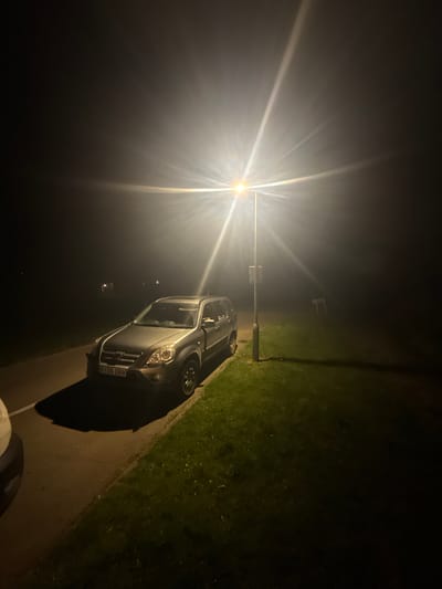 A nighttime scene in Mid Suffolk, United Kingdom. A silver Vauxhall Meriva (license plate KX56 OBB) is parked on a paved area next to a grassy verge. A street light illuminates the scene, creating a strong glare and long shadow. The background is dark. A white moped is in the bottom right corner.