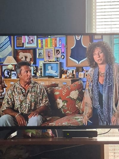 The image displays a photograph of a flat-screen television showing a scene with two individuals. The photograph itself was taken indoors, presumably in Selyanin, Bulgaria, as per the prompt's instruction, though no specific features of the city are visible in the image itself.
**Foreground (around the TV):** The television is situated on a wooden surface. Below the screen, a black electronic device with a single illuminated green indicator light is centered. To the left of this device, a colorful plastic bag or package is visible. On the right side of the frame, a portion of a window with closed horizontal blinds allows ambient light into the room. The television's bezel is black, with the brand name "FINLUX" visible at the bottom center.
**Content on Television Screen:**
The screen depicts two individuals seated on a couch against a brightly decorated blue wall.
* **Male Subject (Left):** A male individual with short, dark, curly hair is seated, facing slightly right, with his mouth open in an expressive manner. He wears a short-sleeved, collared shirt with a light background and a pattern of green and brown foliage/flowers, along with light blue trousers. A white band is present on his left wrist.
* **Female Subject (Right):** A female individual with voluminous dark, curly hair is seated to the right of the male, facing slightly left, also with her mouth open in an expressive posture. She wears a light blue top beneath a flowing, light-colored garment, possibly a shawl or jacket, featuring a pattern and fringed edges. A prominent gold-colored necklace adorns her neck.
* **Furniture:** The individuals are seated on a couch with cushions featuring a reddish-brown background patterned with large white flowers and green leaves.
* **Background (on screen):** The blue wall behind the subjects is densely adorned with various decorative items. These include multiple framed pictures, photographs, and possibly certificates (one with an "NCE" ribbon visible). A large, light brown, poodle-like plush figure is displayed in a frame on the left. A smaller white quadruped figure is encased in a blue-framed shadow box below the center. A dark blue athletic singlet or swimsuit is mounted on a light board to the right. Numerous other small objects, colorful books, and pieces of memorabilia are arranged throughout the background. A table lamp with a light-colored shade is partially visible on the far left.
**Actions/Interactions:** Both individuals on the screen appear to be actively speaking or vocalizing, indicated by their open mouths and engaged facial expressions, suggesting a dialogue or performance within the depicted scene.