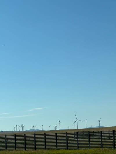 The image depicts a vast, open landscape under a clear blue sky. In the foreground, a weathered wooden fence with horizontal planks stretches across the frame. Behind the fence, a grassy field leads to a distant line of rolling hills. Silhouetted against the sky on the horizon are numerous wind turbines, characteristic of a wind farm. The scene suggests an industrial area integrated into a rural or semi-rural environment. The location is identified as Ereymentaw, Kazakhstan. There are no people visible in the image, and the wind turbines are stationary, indicating no current activity related to them. The environment is arid and expansive.