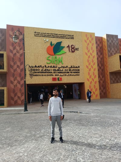 A full-body shot captures a man smiling at the camera, standing on a cobblestone plaza in front of a large, decorative building facade. He wears a grey Calvin Klein sweatshirt, matching grey sweatpants, and black sneakers. His hands are at his sides. The building, likely an exhibition hall, features a beige paneling adorned with vertical wheat stalk patterns in warm brown and orange tones.
Dominating the facade is a large, colorful logo for "SIAM," depicting a fruit (possibly an apple or orange) with green leaves. Above the logo, text in Arabic and French reads: "تحت الرعاية السامية لصاحب الجلالة الملك محمد السادس نصره الله / SOUS LE HAUT PATRONAGE DE SA MAJESTÉ LE ROI MOHAMMED VI QUE DIEU L'ASSISTE." To the right of the logo, "18" and "الدورة / Édition" indicate the 18th edition of an event. Below the logo, more text identifies the event as "الملتقى الدولى للفلاحة بالمغرب / SALON INTERNATIONAL DE L'AGRICULTURE AU MAROC" and specifies the dates and location: "مكناس من 20 إلى 28 أبريل 2026 / MEKNES DU 20 AU 28 AVRIL 2026." Moroccan and Portuguese flags are visible beneath this text.
Several other individuals are seen in the background, moving in and out of the building's entrance. A vintage-style street lamp stands to the left of the central figure. The scene is brightly lit by daylight under a clear sky, casting shadows on the cobblestone ground. The overall setting is the outdoor entrance to the International Agricultural Show in Meknes, Al Machouar – Stinia, Morocco.