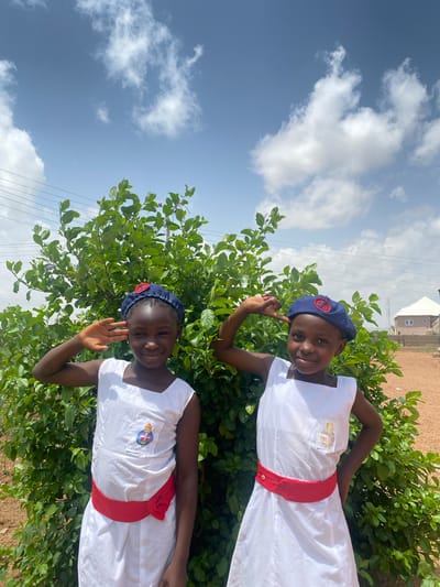 Two young girls are standing in front of a leafy green bush under a partly cloudy sky. The girls are both wearing white dresses with red sashes and blue hats with a badge on the front. The girl on the bottom is waving with her right hand, and the girl on the top is holding up her right index finger. The scene appears to be outdoors in Turu, Nigeria, based on the landscape and foliage. A building is visible in the background.