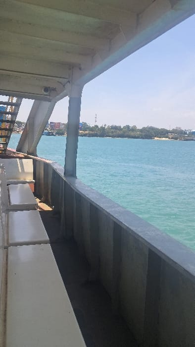 The image depicts a static scene captured from the interior, covered deck of a large vessel or structure, likely a ferry, positioned on turquoise-blue water under a clear sky. In the immediate foreground to the left, a portion of a white, beamed ceiling structure is visible, supported by a vertical grey column. Below the ceiling, two parallel, light-colored benches or seating areas extend horizontally along the left edge of the frame. A dark grey metal railing, composed of vertical panels and a horizontal top rail, forms the edge of the vessel, separating the interior from the body of water.
The midground features a wide expanse of calm, brilliant turquoise water, indicative of a channel or bay under bright sunlight. In the background, a distant coastline is visible, characterized by a mix of natural vegetation, including dense green foliage and sandy areas, and urban development. Prominent features on the coastline include a reddish-brown multi-story building with white accents, alongside other scattered structures. To the far right, a tall communications tower or mast rises above the tree line, and what appears to be port or industrial infrastructure is visible along the water's edge.
No individuals or active interactions are discernible in the scene. The bright, high-angle sunlight suggests a midday setting. This scene is consistent with a view from the Likoni Ferry channel, operating within or adjacent to the Ganjoni/Shimanzi wards in Mombasa, Kenya, with the distant shoreline representing either parts of Mombasa Island or the mainland across the channel.