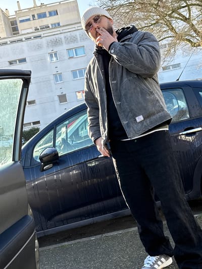 An adult male is depicted standing outdoors on a paved surface in Hérouville-Saint-Clair, France, under clear daylight conditions. The man, facing slightly right and looking towards the viewer, wears a light-colored beanie, round sunglasses, a dark grey or faded black work-style jacket over a black hooded sweatshirt, black trousers, and grey-and-white striped athletic shoes. He possesses a beard and holds a lit cigarette to his mouth with his right hand. His left hand, holding a dark object resembling a smartphone, is partially obscured. To the man's right, a dark blue hatchback or compact MPV vehicle, exhibiting vertical water streaks on its side panels, is parked. An open dark-colored car door is visible in the immediate foreground, positioned to the left. The background features a multi-story, light-toned concrete residential building with numerous windows, and bare tree branches against a clear sky.