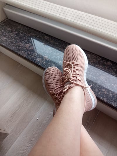 A close-up, high-angle shot shows a person's legs and feet clad in pink knit athletic shoes. The shoes are laced with pink laces and have white soles. The legs are crossed, with one shoe slightly overlapping the other. The shoes rest on a light-colored wooden floor. In the background, a dark, speckled granite surface runs horizontally, and above that, a cream-colored blind with horizontal slatted lines is visible. The scene appears to be indoors, possibly near a window or balcony. There is no indication of the location being in Ankara, Turkey.