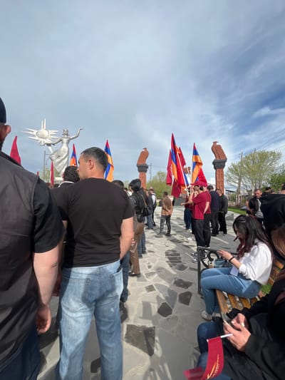 A low-angle shot captures a crowd of people gathered at the Armenian Genocide Memorial in Akhuryan, Armenia. The memorial is adorned with statues, and the Armenian flag is prominently displayed. Individuals are seen standing, sitting on benches, and holding flags. The sky is partly cloudy, and the scene suggests a commemorative event. The ground is paved with stones.