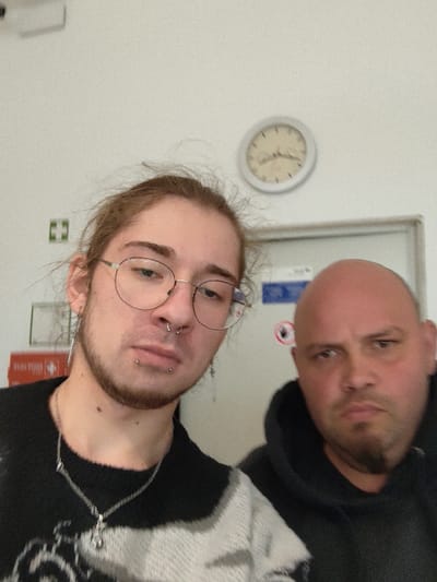 A selfie photograph depicts two males indoors, located in Wiesmoor, Germany. The subject on the left, a young adult, has light brown hair, wears circular metal-rimmed glasses, a septum piercing, two labret piercings, and two elongated earrings. He has visible facial hair. He is wearing a black and white patterned knit sweater and a silver chain necklace with a pendant. His expression is neutral. The subject on the right, an adult male, has a bald head and stubble on his chin. He is wearing a dark hooded garment. His expression is neutral. The background features a light-colored wall. A white circular wall clock, showing approximately 10:45, is mounted in the upper right. To the left of a light-colored door, a green square first-aid sign with a white cross and an 'H' (DIN EN ISO 7010-E003) is visible, along with a small white rectangular object below it. On the door, a blue rectangular sign and a partially visible white circular prohibition sign with a red border and a black hand symbol are present.