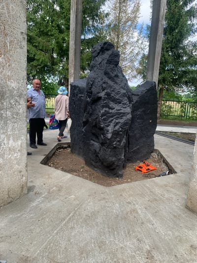 In Arbuzov-Baran, Russia, an outdoor scene depicts a monument under construction. A large, rough-hewn black stone sculpture, possibly a memorial, dominates the center of the frame. It rests on a patch of exposed earth within a concrete foundation that is still being finished. To the left, two individuals stand near the monument. An older man in a checkered blue and white shirt and black pants is looking towards the camera, holding a mobile phone in his right hand. Beside him, a person wearing a light-colored top and a patterned blue and white hat is walking away from the camera, carrying a bag. In the foreground, construction tools are visible, including an orange toolbox with a drill and a circular saw with a power cord. The monument is surrounded by concrete, with some areas appearing rough and unfinished. In the background, lush green trees and a wire fence can be seen. The sky is partly cloudy.