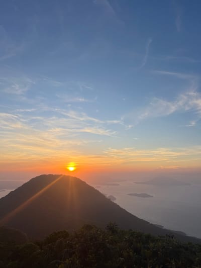 The image depicts a serene sunrise over a mountainous coastal landscape. In the foreground, lush green foliage casts shadows, hinting at a vantage point from a higher elevation. Dominating the mid-ground is a silhouetted mountain peak, its dark contours sharply defined against the vibrant sky. The sun, a brilliant orb of light, is positioned just above the mountain's summit, casting long, golden rays that stretch across the scene, illuminating the sky with hues of orange, yellow, and soft pink. Wispy clouds streak across the horizon, adding texture to the dawn sky, which transitions into a clear blue overhead.

The background reveals a calm body of water, likely a bay or ocean, dotted with several islands of varying sizes. The water appears still, reflecting the soft light of the sunrise, and is partially obscured by a gentle haze that softens the distant view. This scene is consistent with the geography of the area surrounding El Cacao, in the department of Santa Ana, El Salvador, known for its volcanic landscapes and proximity to Lake Coatepeque, which features islands and is often surrounded by mist. No people or discernible human activity are visible in the image.