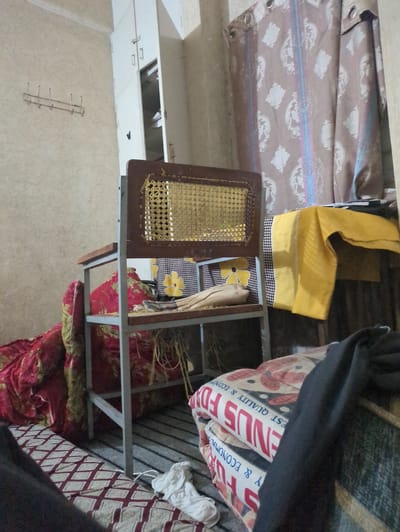 The image depicts a cluttered indoor scene, possibly a room in a home or a storage area, in Abbottabad, Pakistan. The overall impression is one of disarray, with various items piled and spread out.
Dominating the foreground and midground is a metallic frame, likely part of a bed or a shelving unit, which is filled with various textiles. A red blanket or quilt with a floral pattern is prominently displayed, folded and draped over parts of the frame. To the right of this frame, more bedding with a diamond pattern is visible. A dark piece of fabric, possibly clothing or a larger textile, is draped over the top of the metallic structure, obscuring some of the items beneath.
In the midground, a small wooden-framed piece with a woven cane insert is positioned. Inside this frame, a yellow cloth with a black and white checkered border is visible, along with a floral-patterned fabric with a yellow flower.
The background reveals more textiles, including what appears to be a rolled-up mattress or bedding with text printed on it. The text is partially obscured but includes "EST QUALITY & ECONOM" and "ENDUS" which might be part of a brand name or product description. Above the metallic frame, there are stacks of what look like folded blankets or sheets with a patterned design, featuring a light brown background with white motifs.
The setting appears to be an interior space, with a wall visible on the left, painted in a light, textured color. Towards the top, a section of a ceiling or a higher surface is visible, with some sort of opening or recessed area. There is also a metallic rack with several hooks mounted on the wall. The lighting suggests it is daytime, but the overall lack of clear light sources and the clutter make it difficult to ascertain the exact time of day or weather. No people are visible in the image. The general atmosphere is one of disorganization and perhaps the storage of household items.