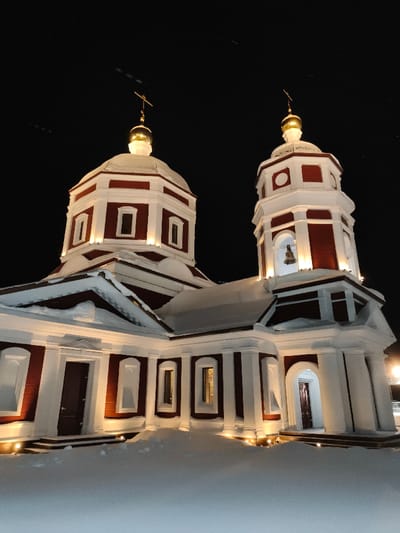 The image displays an Orthodox church or chapel at night, situated in the city of Im. Gastello, Russia. The structure is characterized by deep red walls and contrasting white architectural elements, including cornices, pilasters, and window frames. It features a central octagonal dome topped with a golden cupola and a cross, and an adjacent octagonal bell tower, also crowned with a golden cupola and cross. A single bell is observable within the bell tower's upper archway. The building's exterior is illuminated by multiple warm yellow uplights positioned at ground level and along architectural ledges. The ground, roofs, and ledges are covered in a substantial layer of snow, indicative of winter conditions. The sky above is dark. No individuals, actions, or interactions are depicted.