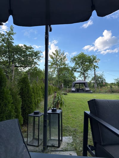 The image shows an outdoor patio area, possibly in Lozno, Bulgaria. In the foreground, a portion of a dark-colored outdoor sofa is visible. To the left, two black metal and glass lanterns sit on a stone surface. A tall, dark umbrella pole is centrally located, extending upwards, with the dark fabric of the umbrella visible at the top of the frame. Behind the lanterns, a line of manicured green evergreen bushes forms a hedge. A wooden fence with stone posts runs along the edge of the property behind the bushes. The middle ground features a neatly mown green lawn, with a dark gazebo structure with seating and a table located further back. Several trees are visible throughout the yard, and the sky above is blue with scattered white clouds. The overall impression is a well-maintained outdoor living space.