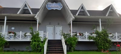 The image shows the facade of a gray, wooden, two-story building with multiple triangular gables and a dark shingle roof. A white balcony with white railings runs along the front of the building. Planter boxes with red flowers and greenery are placed on the balcony. Two sets of white double doors are visible on the ground floor, with a set of dark wooden stairs leading up to them in the center. Above the central entrance, a decorative blue sign with white floral accents reads "Townhouse 2". The building is surrounded by lush green grass and vegetation, with red roses visible in the foreground. The overall scene appears to be a residential complex or hotel. The lighting suggests it is daytime, possibly overcast. The location is Ikindzhi Nyugedi, Azerbaijan. There are no people or animals visible in the image.