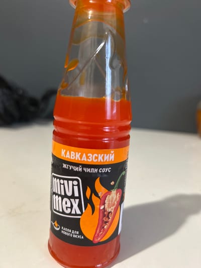 A clear plastic bottle of Mivi Mex chili sauce is displayed on a white surface, casting a shadow to the right. The bottle is filled with a bright orange-red sauce. The label features the brand name "Mivi Mex" in large white letters with a black outline. Above this, in orange text on a black background, are the Russian words "КАВКАЗСКИЙ" and "жгучий чили соус," which translate to "Caucasian" and "hot chili sauce" respectively. A stylized image of a chili pepper and flames is also present on the label. Below the brand name, more Russian text reads "капля для нового вкуса," meaning "a drop for a new taste." The background is a muted gray. The overall scene is indoors and well-lit. There are no people or animals present.