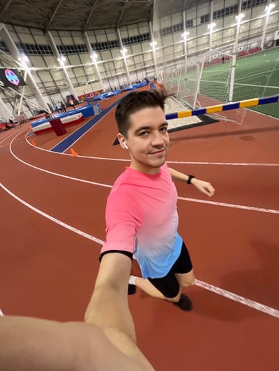 A man takes a selfie indoors at a track and field facility. He wears a pink and blue ombre shirt, black shorts, and black sneakers. An athletic track in shades of brown is visible beneath him. The track has white lines painted on it. Behind the man are rows of seating, hurdles, and a net. The ceiling is high and industrial with numerous lights. He wears a smartwatch and an earbud.