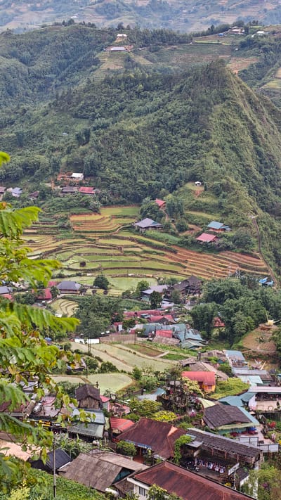 A high-angle shot of Cat Cat Village in Vietnam. Terraced rice paddies cascade down a lush, green hillside interspersed with residential homes. The village proper, in the foreground, reveals buildings with red and green tiled roofs. A few people are seen walking in the village. The landscape is dominated by green vegetation.